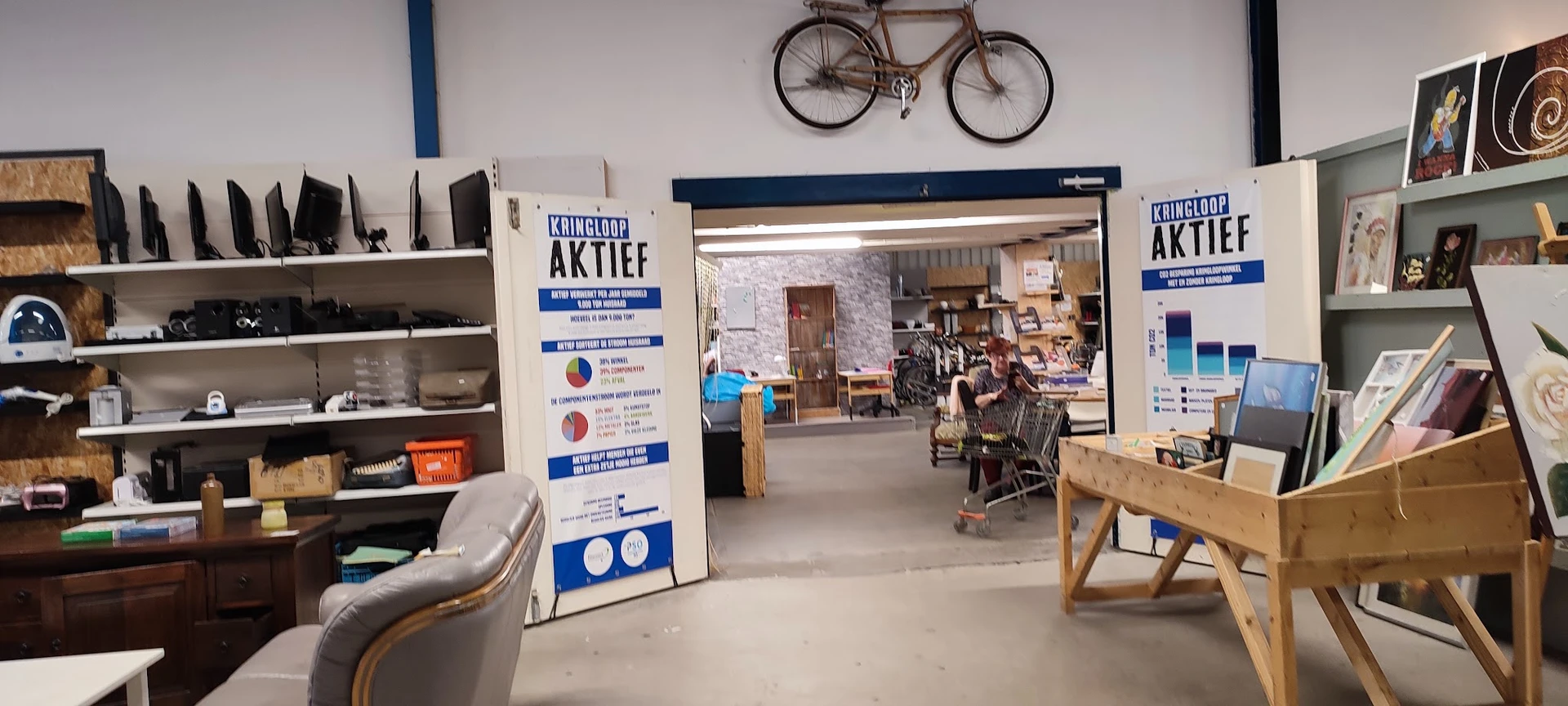 Binnenaanzicht van kringloopwinkel met stellingen, toonbanken, winkelwagen en klanten; fiets aan muur