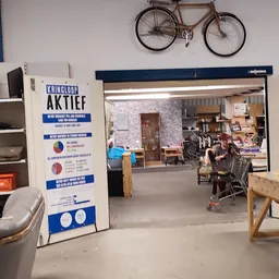 Binnenaanzicht van kringloopwinkel met stellingen, toonbanken, winkelwagen en klanten; fiets aan muur