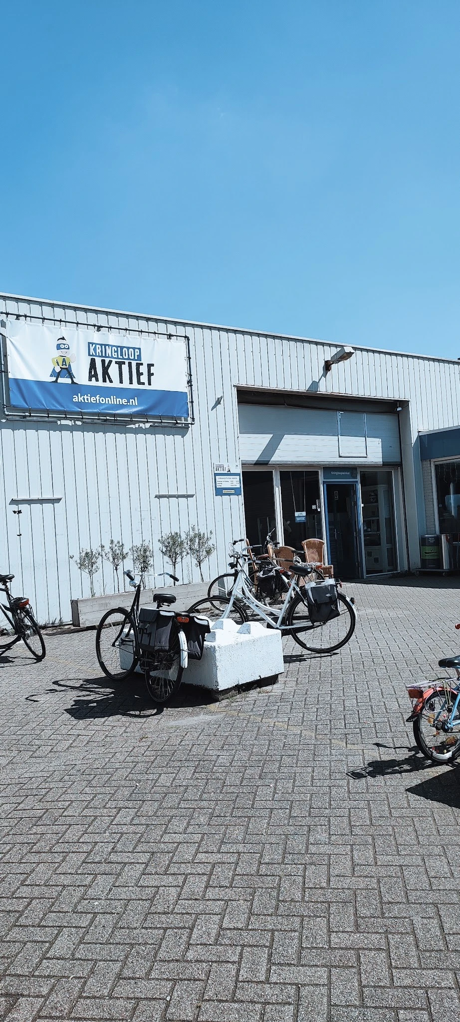 Voorgevel van kringloopwinkel met witte golfplaten wand, ingang en uithangbord, fietsen geparkeerd ervoor