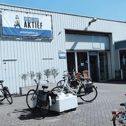 Voorgevel van kringloopwinkel met witte golfplaten wand, ingang en uithangbord, fietsen geparkeerd ervoor
