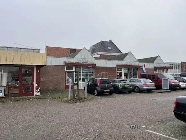 Voorgevel van kringloopwinkel met rode deur, parkeerplaats met auto's, Nederlands vlaggetje en woonhuizen op achtergrond