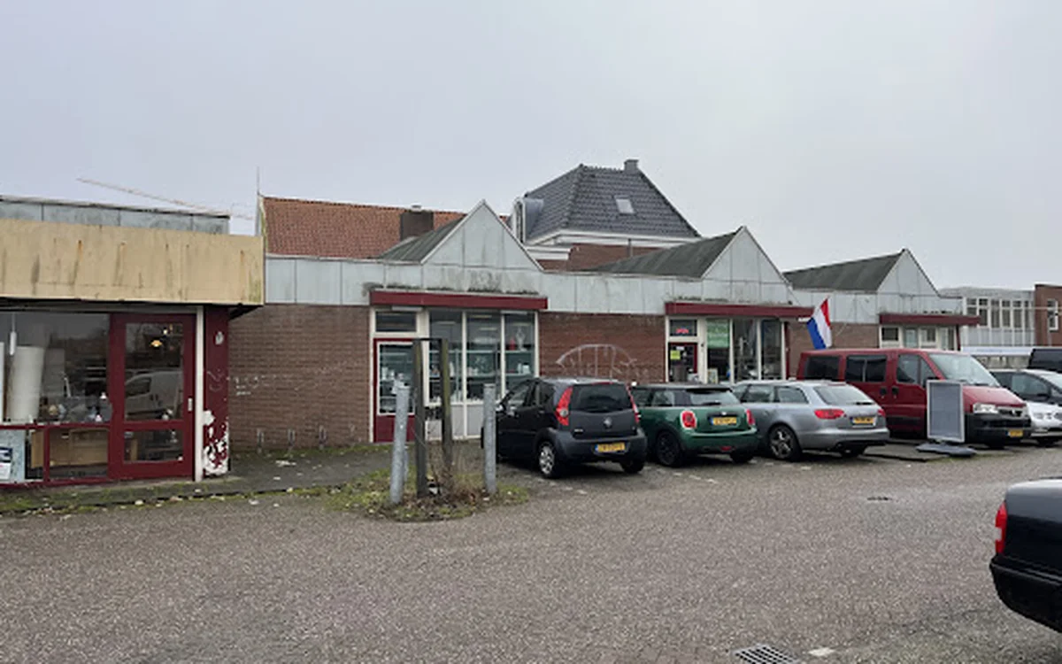 Voorgevel van kringloopwinkel met rode deur, parkeerplaats met auto's, Nederlands vlaggetje en woonhuizen op achtergrond
