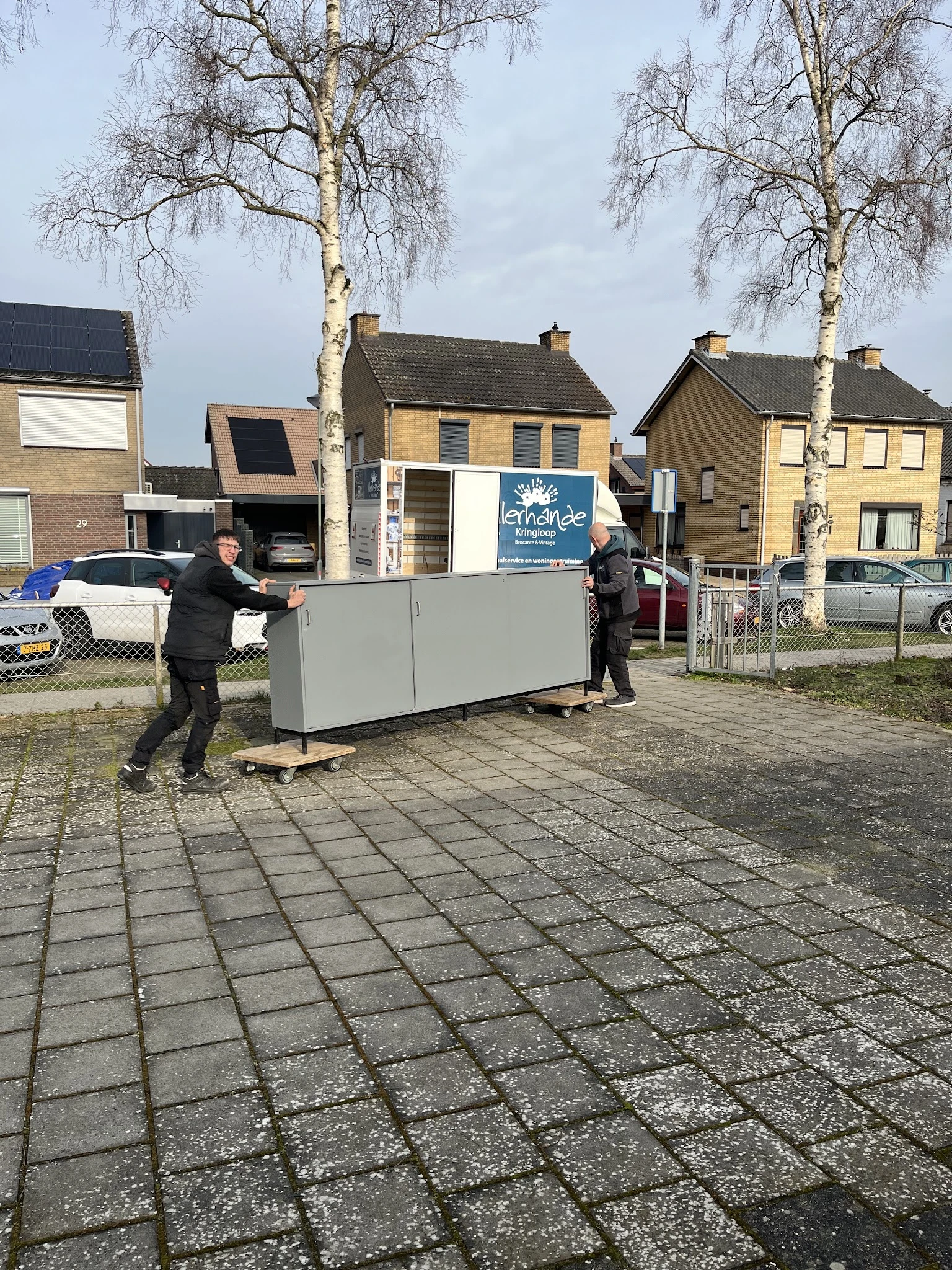 Twee personen verplaatsen een grote grijze kast op een plein voor winkelpanden