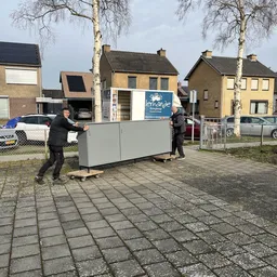 Twee personen verplaatsen een grote grijze kast op een plein voor winkelpanden