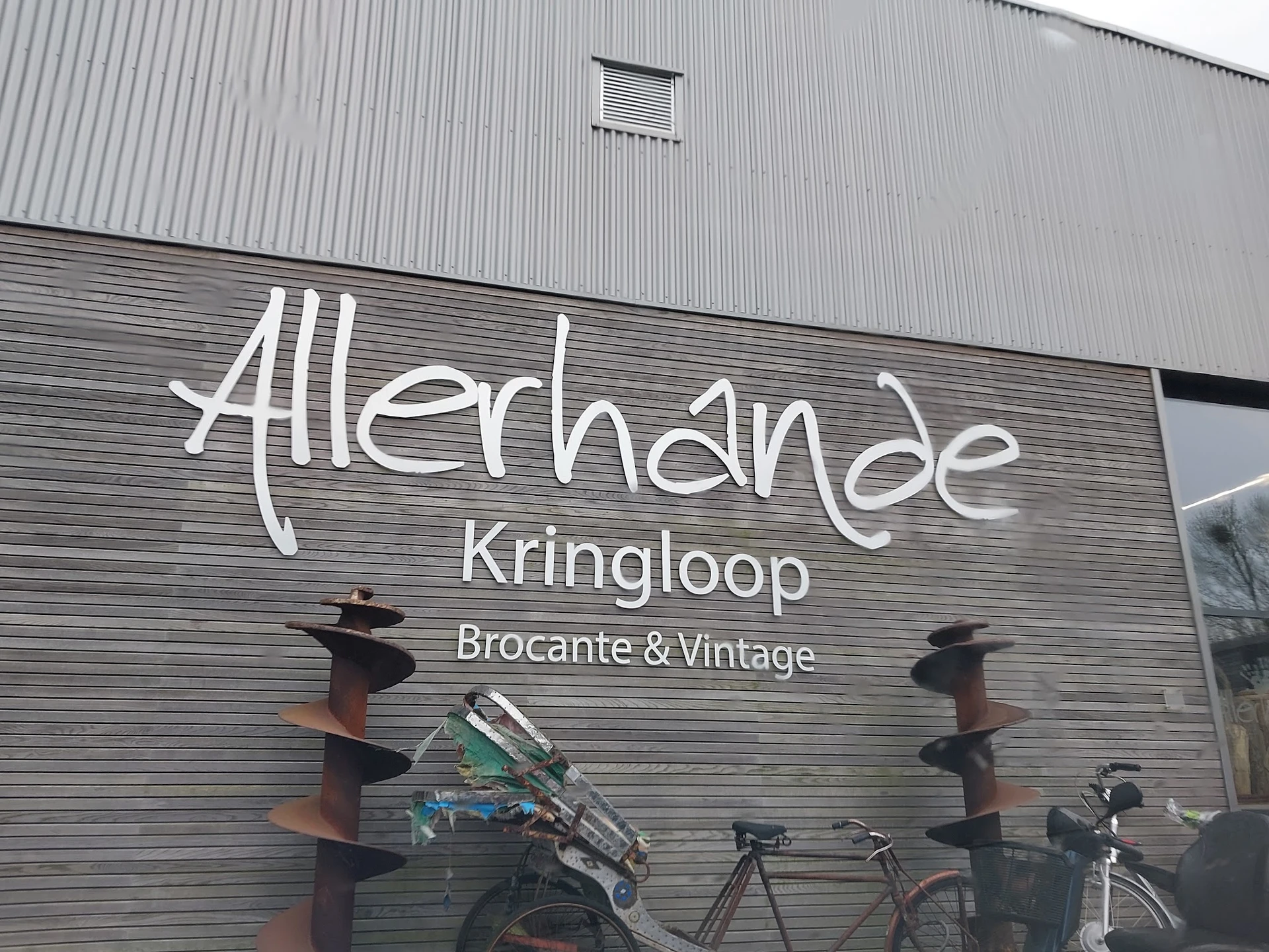 Gevel van kringloopwinkel met wit logo 'Allerhande Kringloop' en ondertitel 'Brocante & Vintage', met daaronder enkele fietse