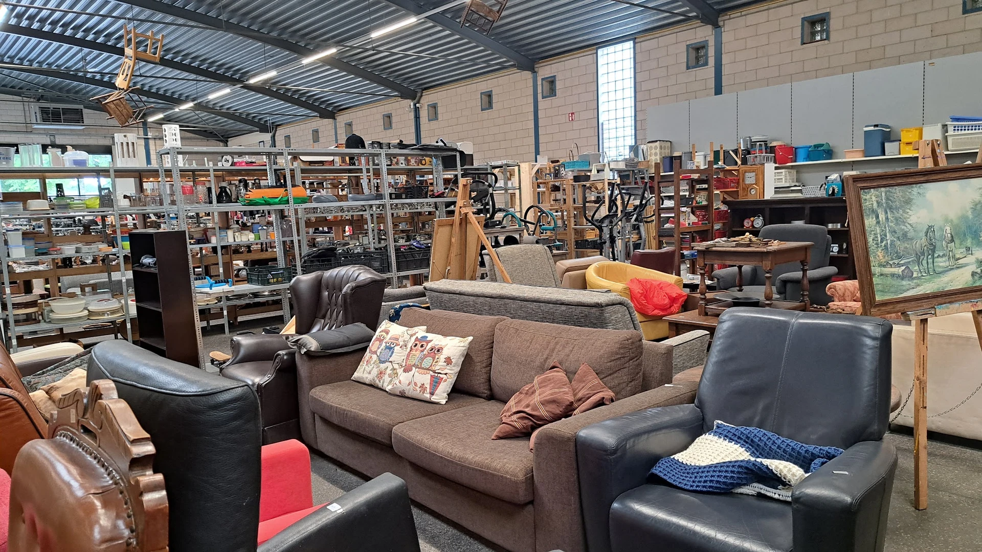 Winkelvloer met diverse meubels: grijze bank, zwarte stoelen, rood kussen, en stellingen met spullen op de achtergrond