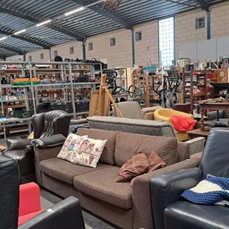 Winkelvloer met diverse meubels: grijze bank, zwarte stoelen, rood kussen, en stellingen met spullen op de achtergrond