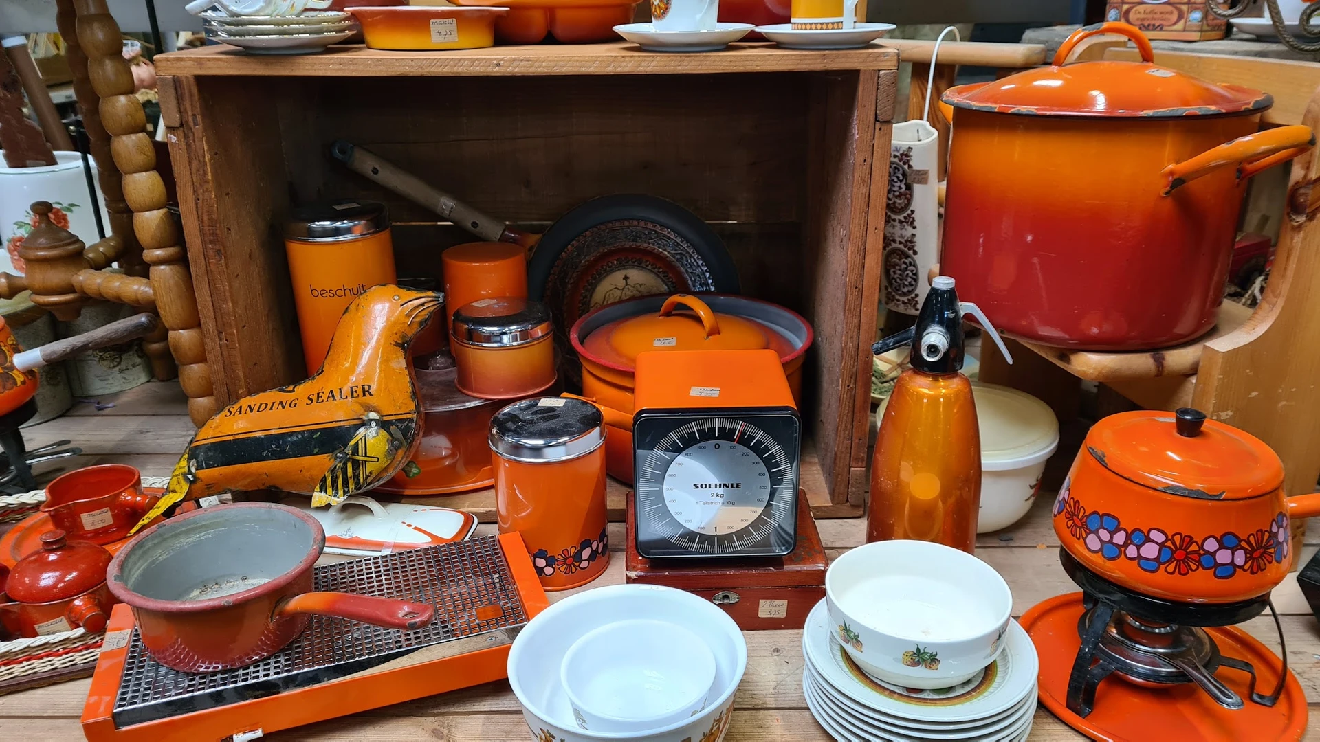 Uitgestalde tweedehands huishoudartikelen: oranje serviesgoed, glazen potten, weegschaal en keukenspullen op en in een houten