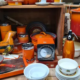Uitgestalde tweedehands huishoudartikelen: oranje serviesgoed, glazen potten, weegschaal en keukenspullen op en in een houten