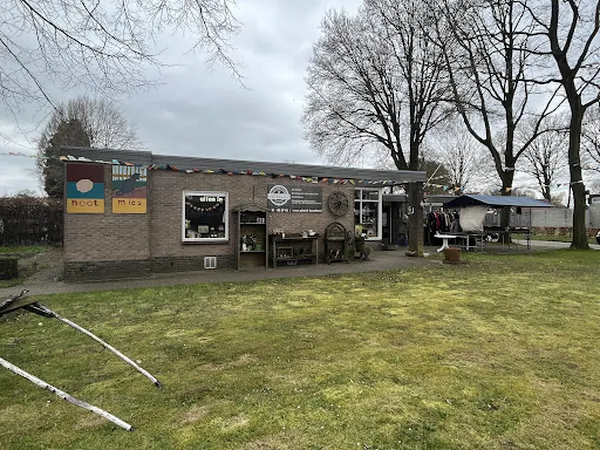 Voorgevel van kringloopwinkel met bakstenen pui, uithangborden en donkere etalageruiten, omgeven door grasveld en kale bomen.