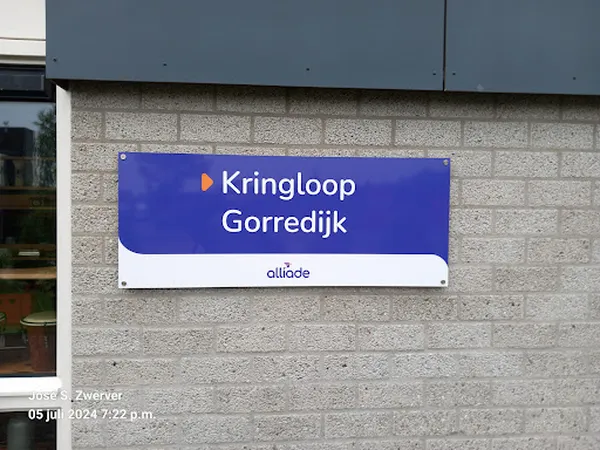 Blauw winkelnaambord 'Kringloop Gorredijk' van Alliade op grijze muur van winkelgebouw