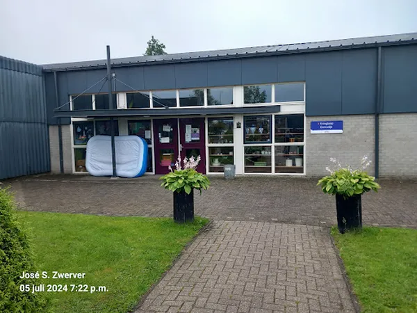 Voorgevel van kringloopwinkel met bakstenen en metalen constructie, grote ramen, bloembakken en doorgezakte luifel