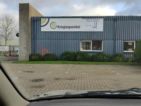 Gevel van kringloopwinkel met grijze golfplaten wand, bakstenen zijkant en witte ramen, winkelnaam zichtbaar op wit bord