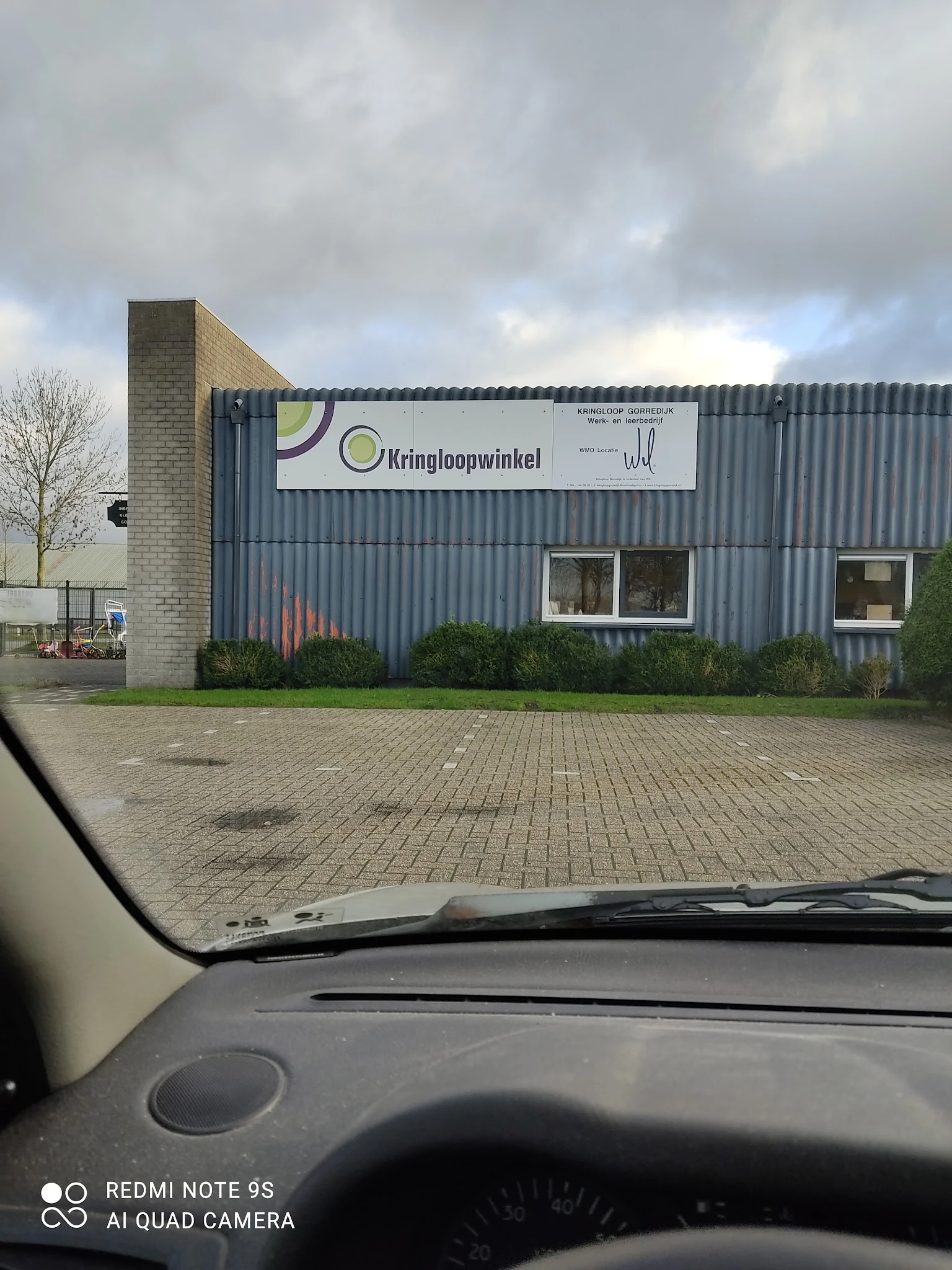 Gevel van kringloopwinkel met grijze golfplaten wand, bakstenen zijkant en witte ramen, winkelnaam zichtbaar op wit bord