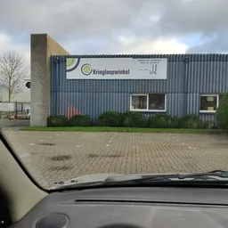 Gevel van kringloopwinkel met grijze golfplaten wand, bakstenen zijkant en witte ramen, winkelnaam zichtbaar op wit bord