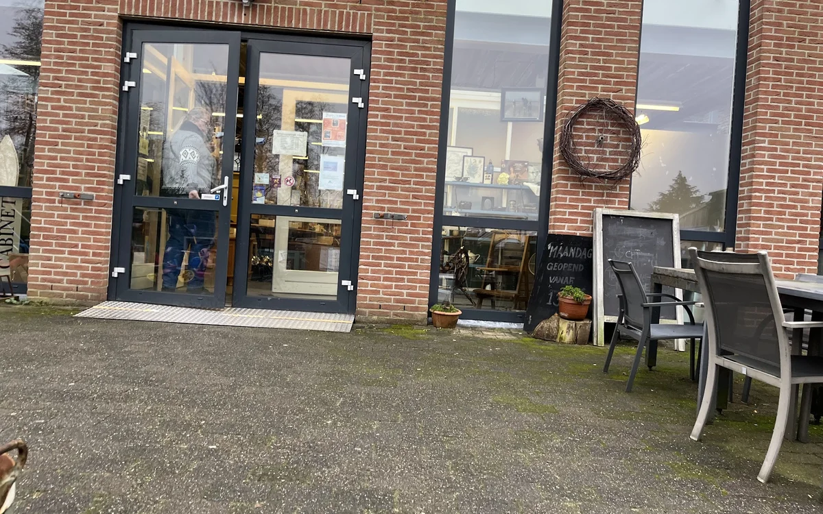 Bakstenen gevel van antiekwinkel met zwarte deur- en raampartijen, terras met stoelen op het plein ervoor