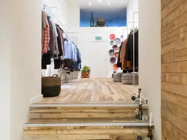 Kledingafdeling met rekken vol tweedehands kleding, plantjes en decoratieve items in een ruime winkelruimte met houten vloer