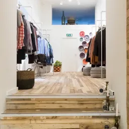Kledingafdeling met rekken vol tweedehands kleding, plantjes en decoratieve items in een ruime winkelruimte met houten vloer