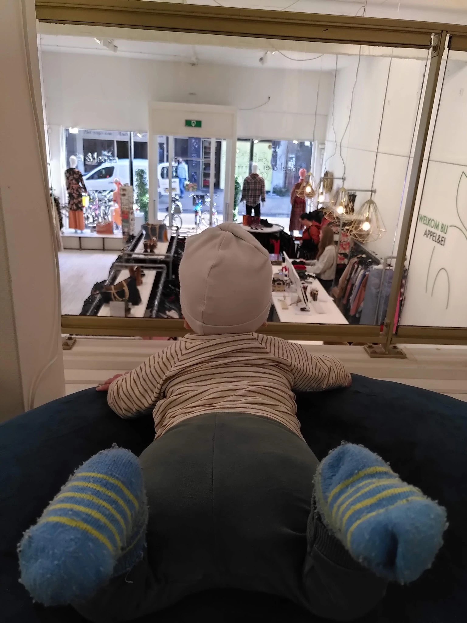 Mannequin met gestreepte trui en grijze broek voor winkelraam met kledingrekken en uitgeklede poppen op achtergrond