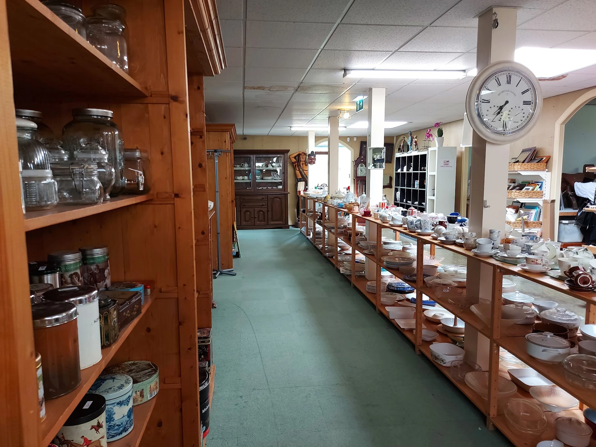 Binnenzicht kringloopwinkel met houten stellingen vol servies en huishoudartikelen, groen linoleum vloer, plafondverlichting 