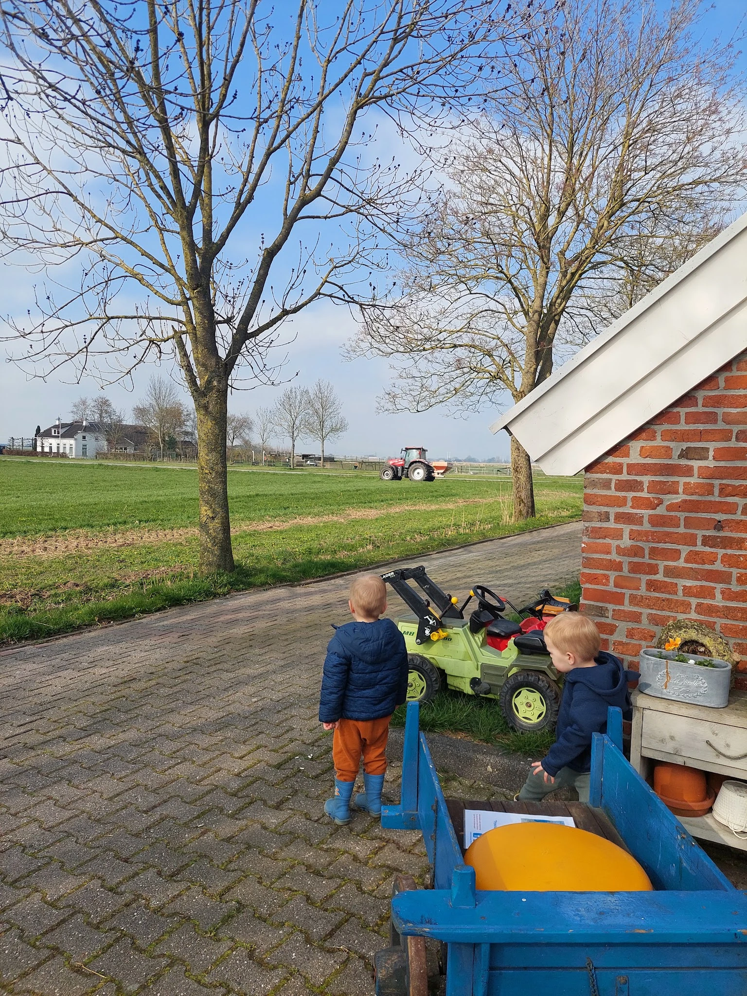Twee kinderen spelen met een gele speelgoedtractor op een terras naast een bakstenen muur, met uitzicht op weiland en een ech