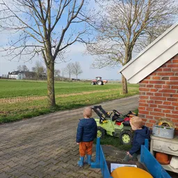 Twee kinderen spelen met een gele speelgoedtractor op een terras naast een bakstenen muur, met uitzicht op weiland en een ech