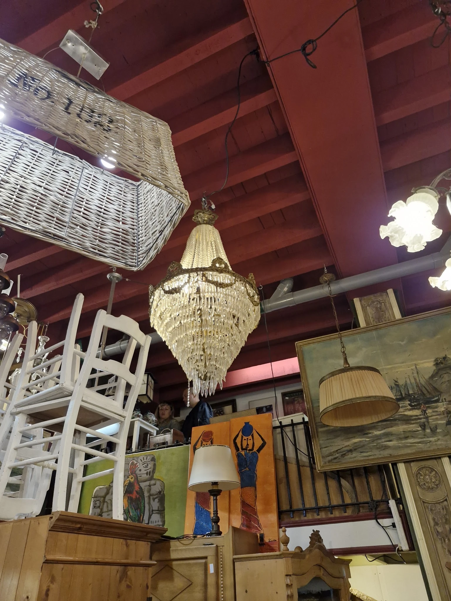 Interieur van kringloopwinkel met kristallen kroonluchter, witte rieten meubels en schilderijen tegen paarse wand
