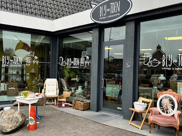 Voorgevel van kringloopwinkel met grote ramen, zwarte kozijnen en ovaal uithangbord. Voor de winkel diverse meubels en voorwe