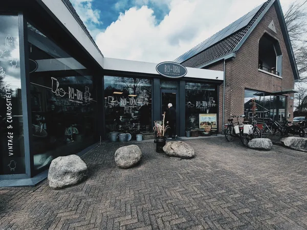 Donkergrijze gevel van kringloopwinkel met grote ramen, bakstenen zijgevel en grote keien voor de ingang