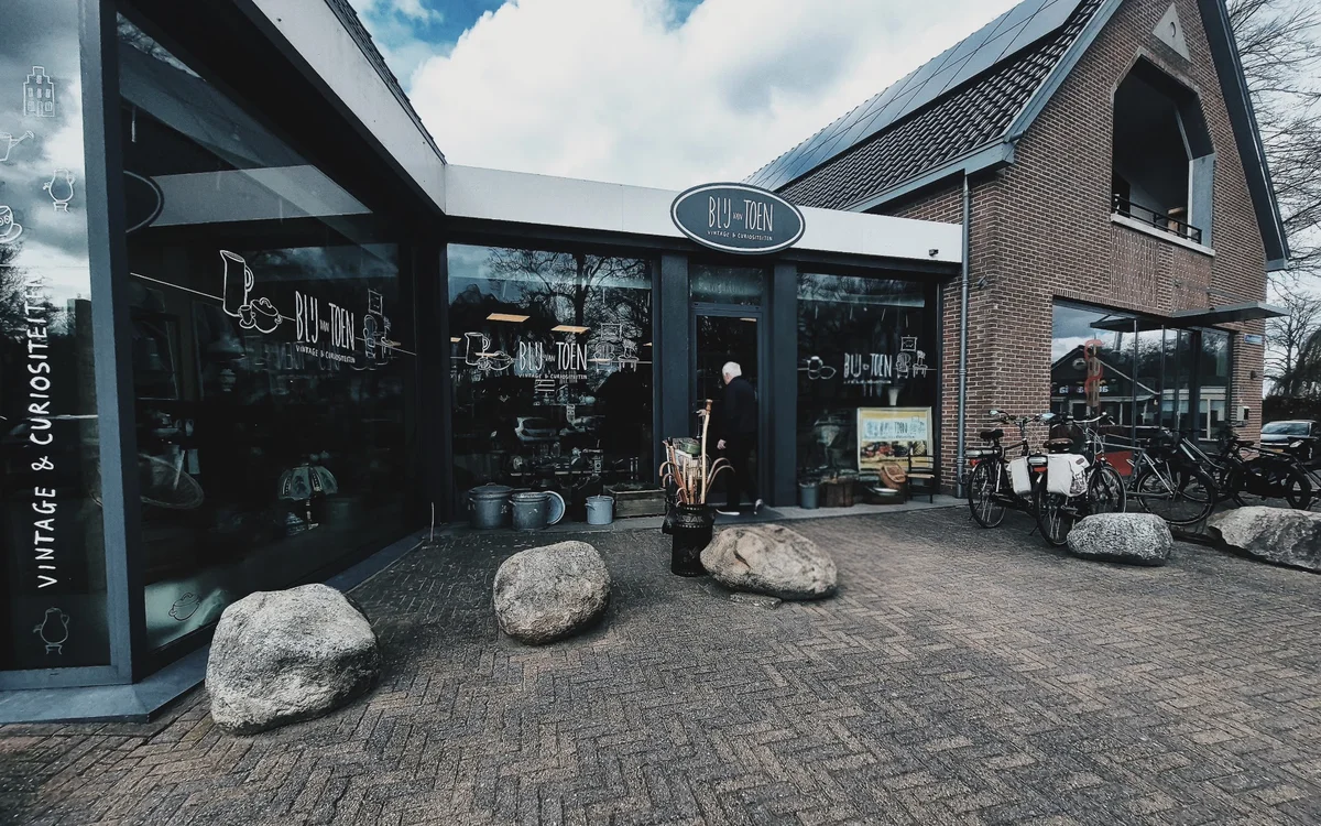 Donkergrijze gevel van kringloopwinkel met grote ramen, bakstenen zijgevel en grote keien voor de ingang