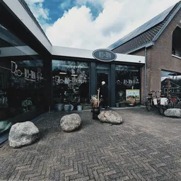 Donkergrijze gevel van kringloopwinkel met grote ramen, bakstenen zijgevel en grote keien voor de ingang