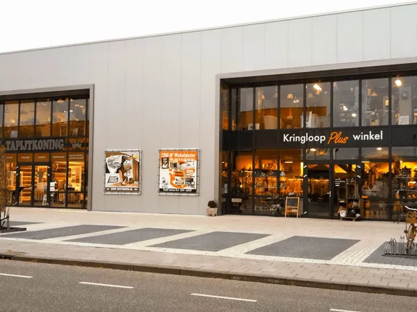 Gevel van kringloopwinkel met twee etalages en donkere ramen, winkelnaam 'Kringloop Plus winkel' zichtbaar