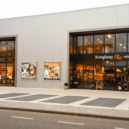 Gevel van kringloopwinkel met twee etalages en donkere ramen, winkelnaam 'Kringloop Plus winkel' zichtbaar