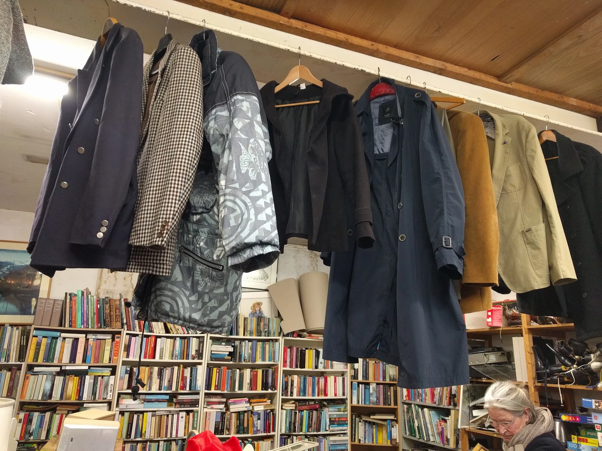Jassen en mantels opgehangen aan een rek boven boekenkasten in een kringloopwinkel