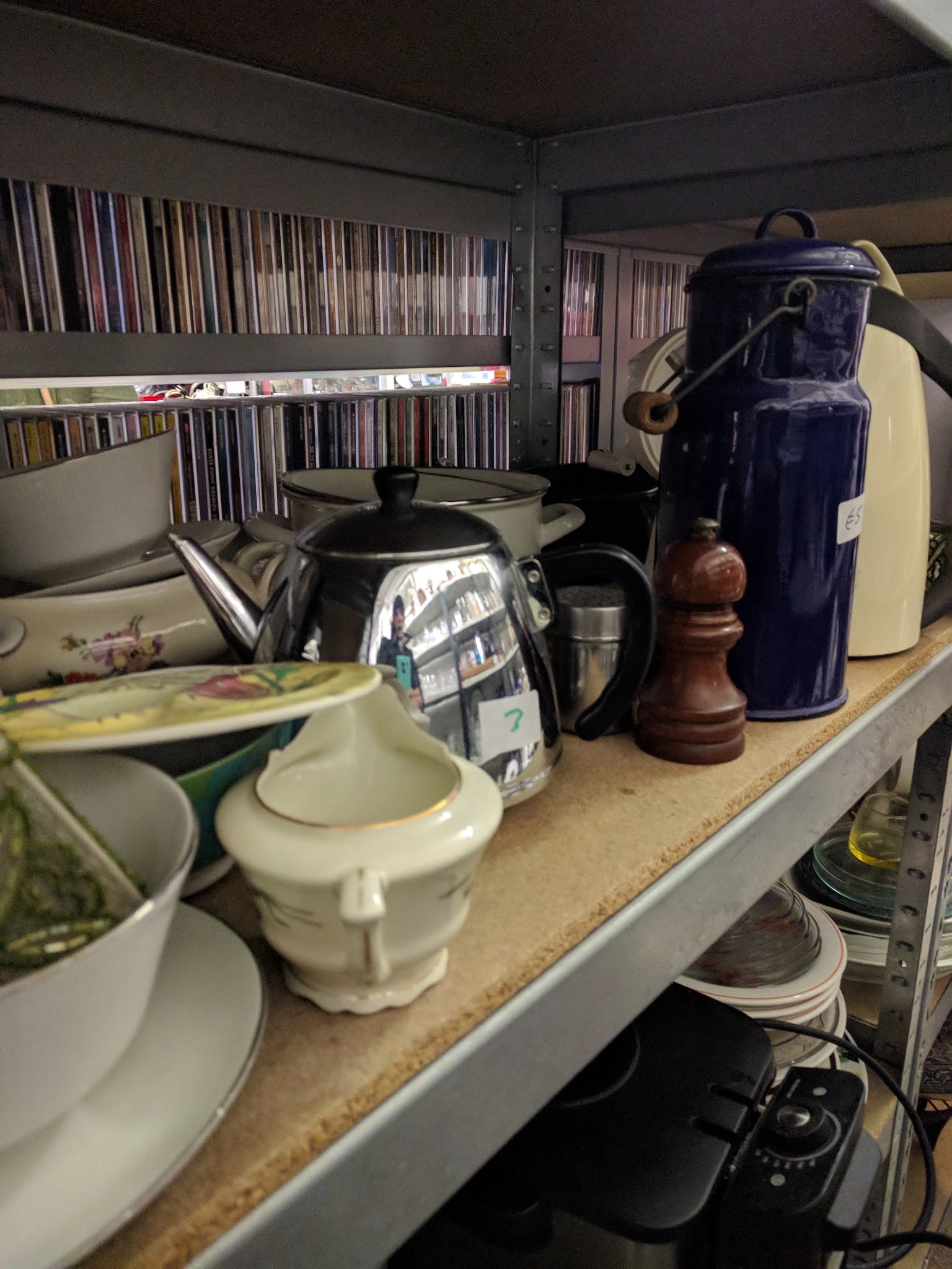Stellingkast met servies, theepotten, karaffen en keukenspullen; op achtergrond cd's