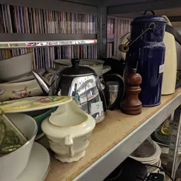 Stellingkast met servies, theepotten, karaffen en keukenspullen; op achtergrond cd's