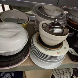 Uitgestalde serviesgoed en pannen op planken, waaronder witte borden, schalen met zilveren deksels en zwarte pannen