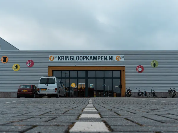 Gevel van kringloopwinkel Kringloopkampen met naam op de pui, grote ramen, ingang en parkeerplaats ervoor