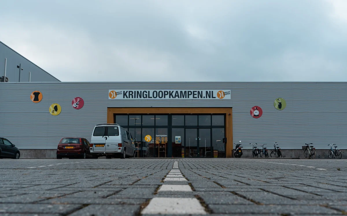Gevel van kringloopwinkel Kringloopkampen met naam op de pui, grote ramen, ingang en parkeerplaats ervoor