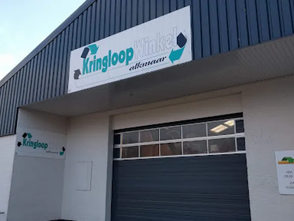Witte gevel van kringloopwinkel met bedrijfsnaam en logo op uithangbord, zwarte garagedeur en raam