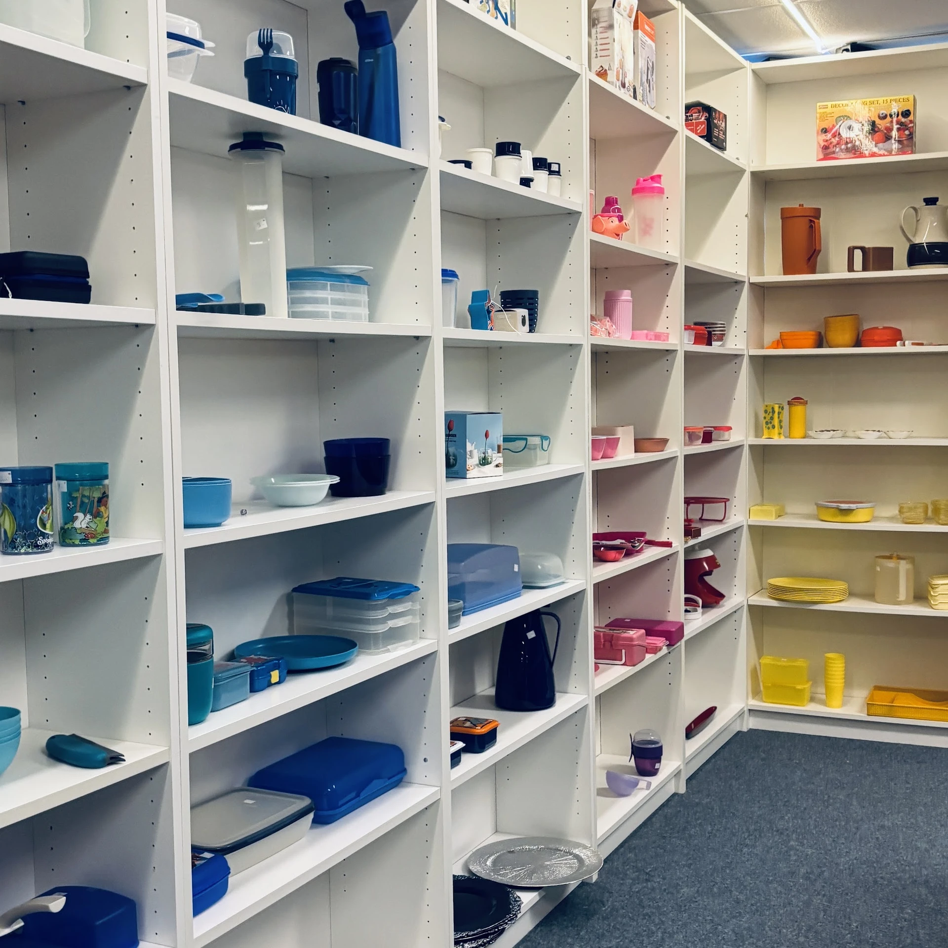 Witte stellingen gevuld met huishoudartikelen in verschillende kleuren: blauwe, roze en gele kunststof bakjes, dozen en servi