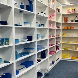 Witte stellingen gevuld met huishoudartikelen in verschillende kleuren: blauwe, roze en gele kunststof bakjes, dozen en servi