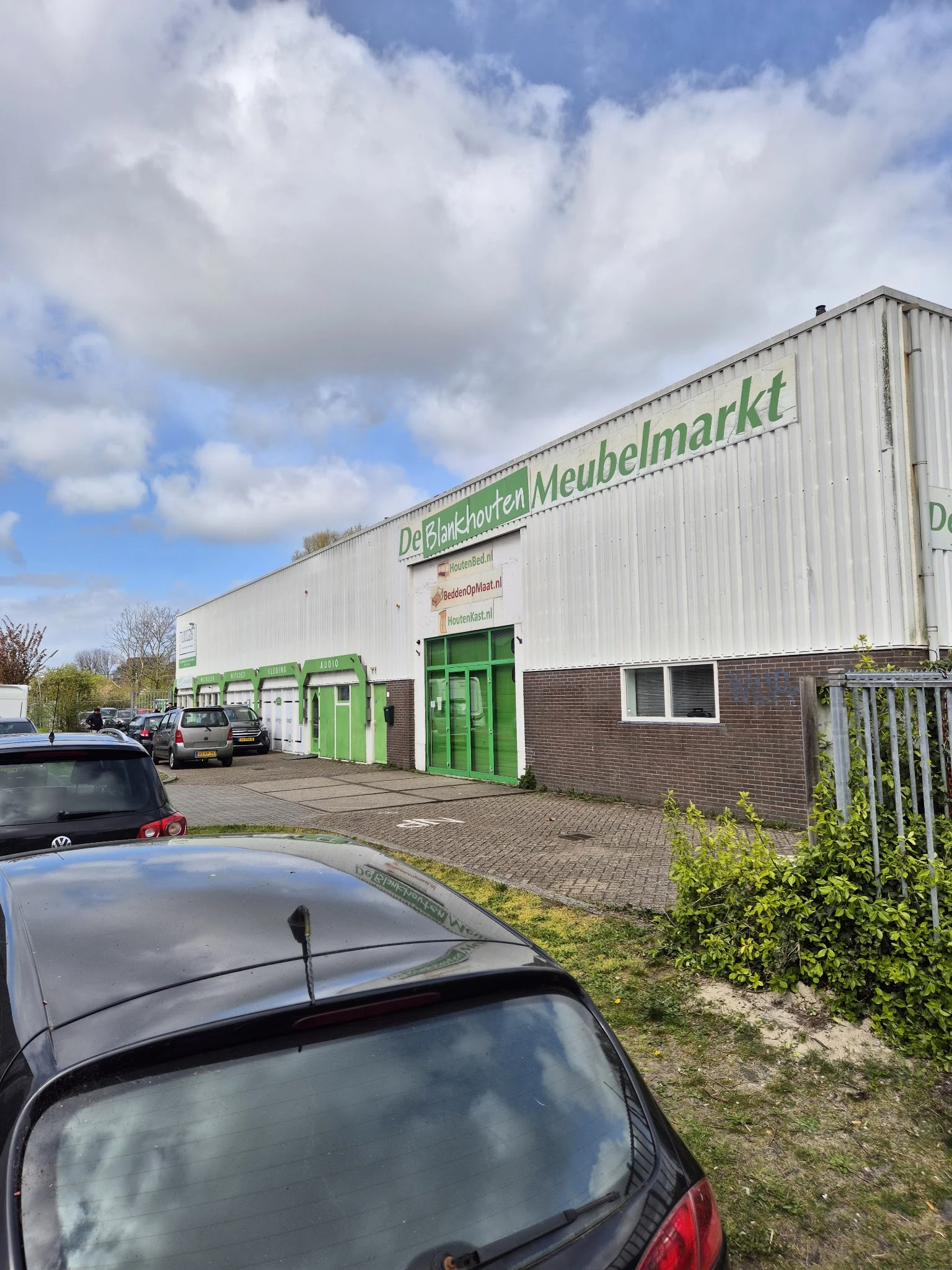Witte gevel van kringloopwinkel met groene deuren en ramen, parkeerplaats voor