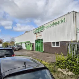 Witte gevel van kringloopwinkel met groene deuren en ramen, parkeerplaats voor