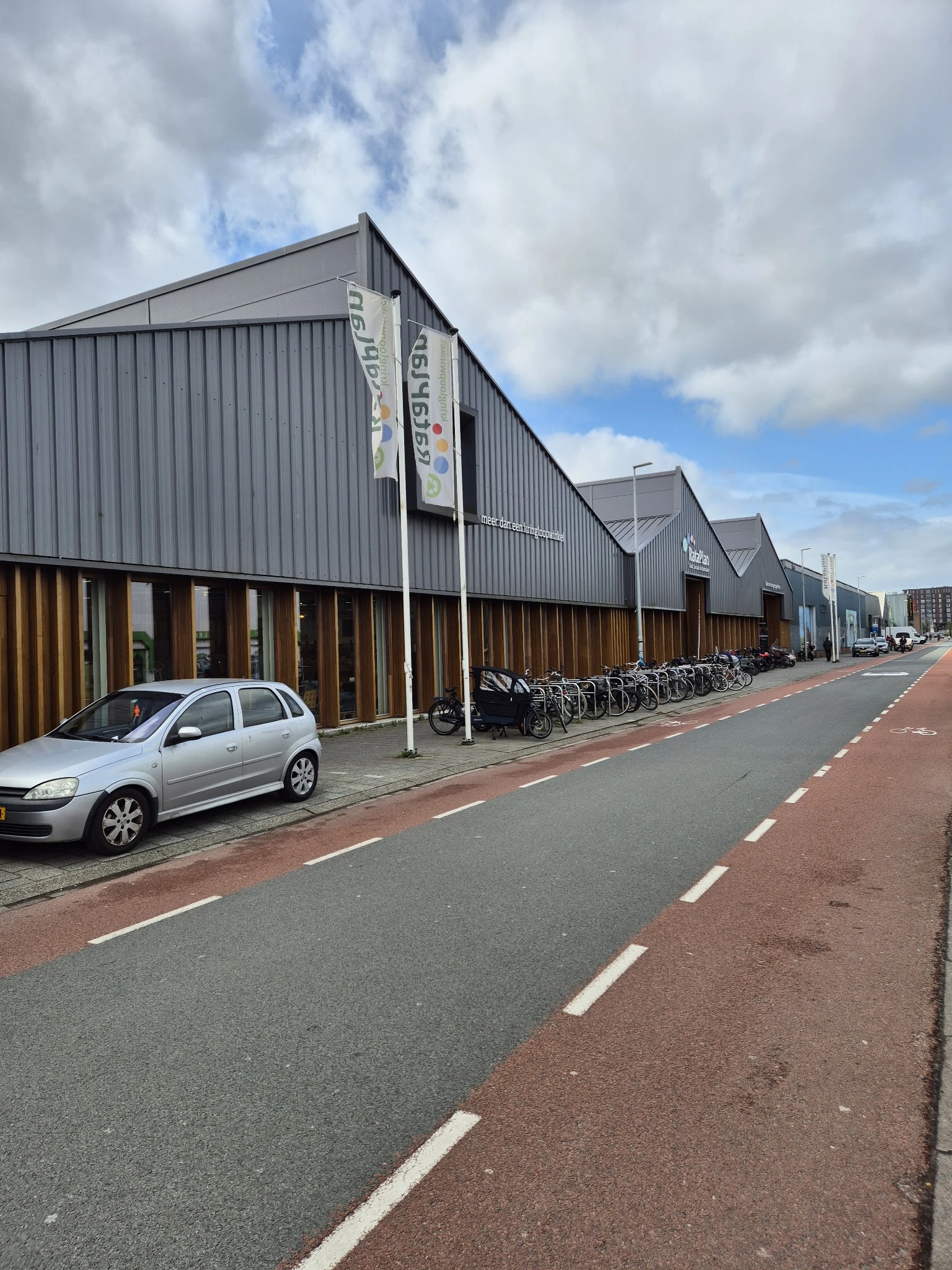 Grijze loods met bruine toegangsdeuren en fietsenstalling langs een rode voetpad