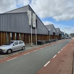Grijze loods met bruine toegangsdeuren en fietsenstalling langs een rode voetpad