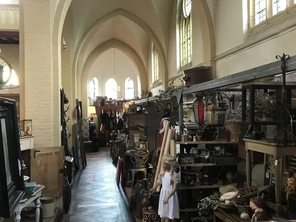 Lange gang in een kerk met hoge gewelven, ingericht als kringloopwinkel met stellingen vol tweedehands spullen aan beide zijd