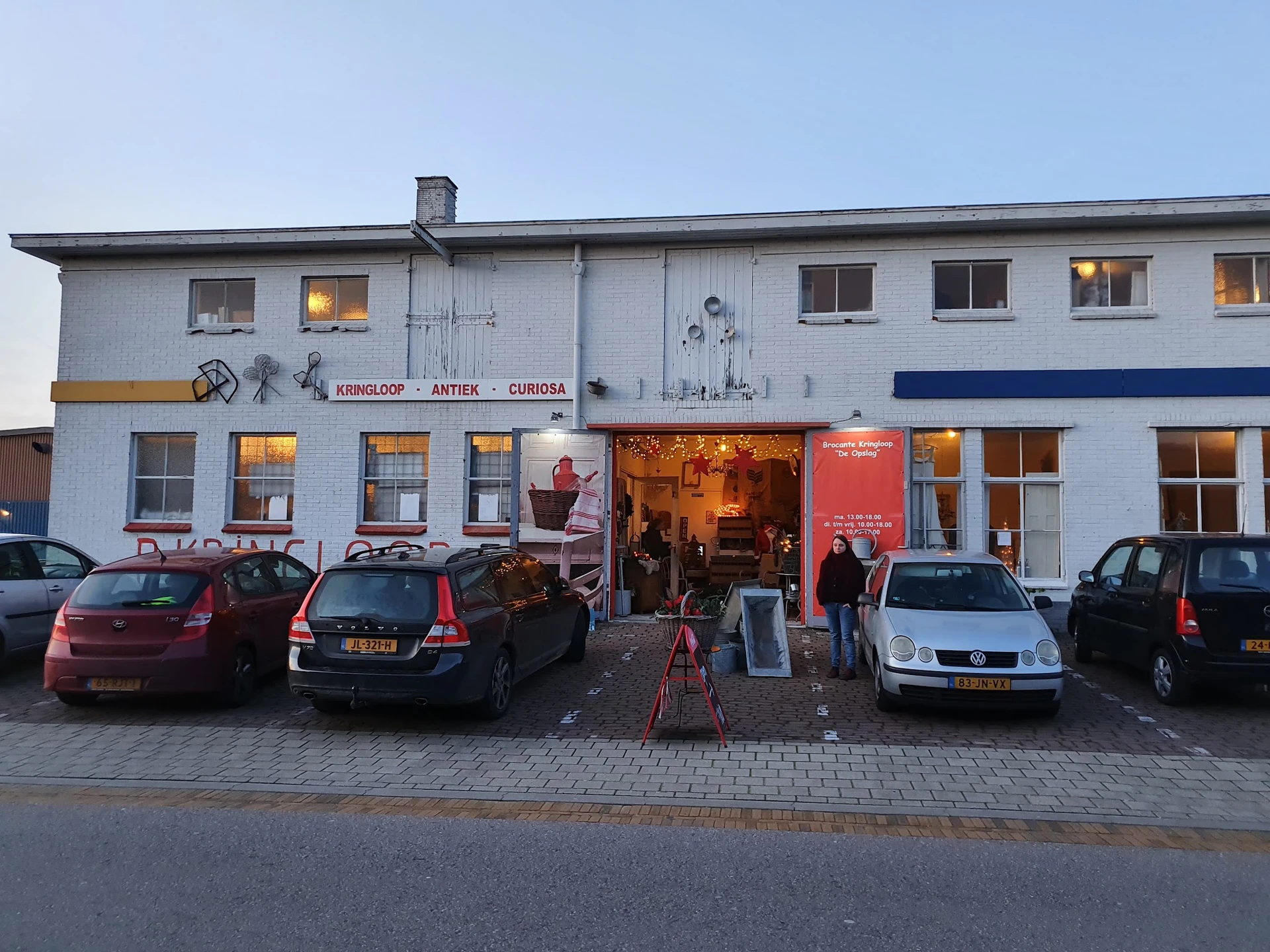 Witte gevel van kringloopwinkel met verlichte etalages en parkeerde auto's ervoor bij schemering