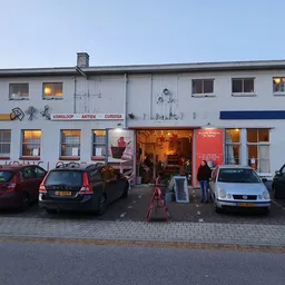 Witte gevel van kringloopwinkel met verlichte etalages en parkeerde auto's ervoor bij schemering