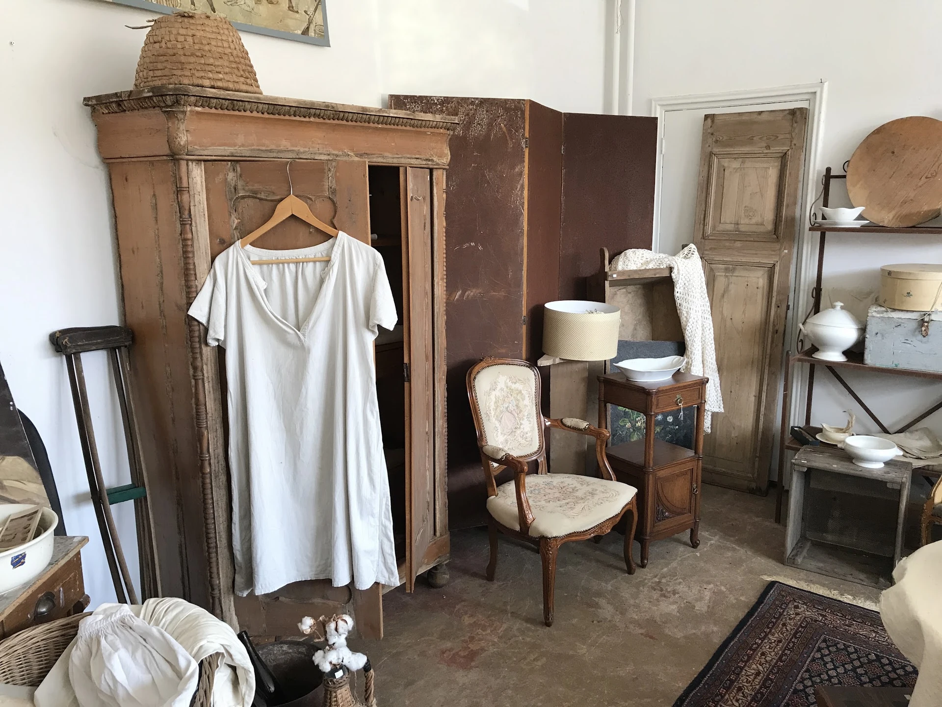 Interieur van een brocantezaak met houten kasten, vintage meubels, een witte jurk op hanger, stoelen en diverse huishoudelijk